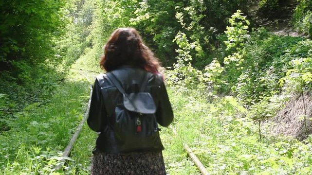 Hiking and nature walk, young woman with backpack walking along overgrown abandoned railway in green forest. Eco tourism concept, woman walks away from camera and looks back