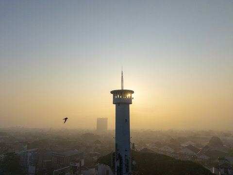 Aerial view of a tall white minaret tower silhouetted against a hazy golden sunset sky with a bird flying nearby over the urban landscape of Malang, East Java, Indonesia.
