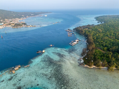 Aerial view of Karimun Jawa island featuring turquoise coral reefs, floating fish farms, and lush tropical forest along the coast of Karimunjawa, Central Java, Indonesia.