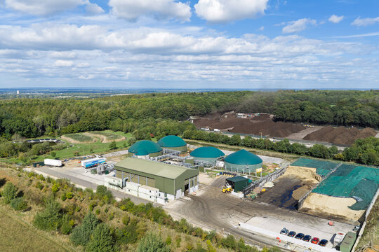 Aerial view of biogas plant with green domed digesters, industrial buildings, and compost piles surrounded by forest in United Kingdom.