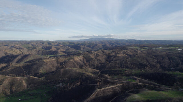 Aerial view of rolling hills near Odeceixe, featuring charred vegetation, patches of green, and distant mountains under a blue sky in Odeceixe, Faro District, Portugal.