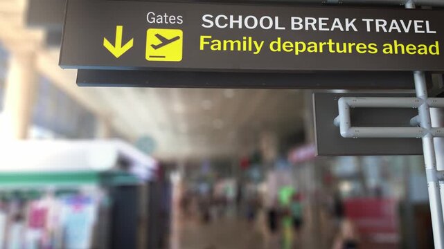 Airport terminal sign indicating directions to the gates for school break travel and family departures, with a blurred background of a busy concourse full of travelers walking towards their flight