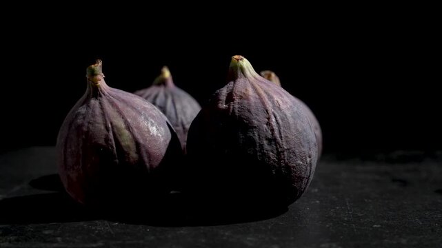 A small group of whole purple figs slowly rotates on a dark stone surface against a black background in moody side lighting.