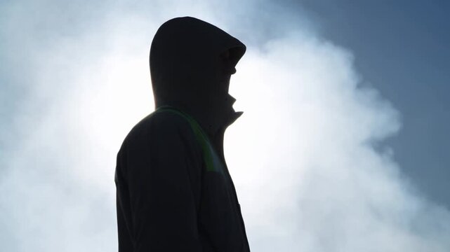Hooded person standing in side profile against a bright cloudy sky with subtle movement