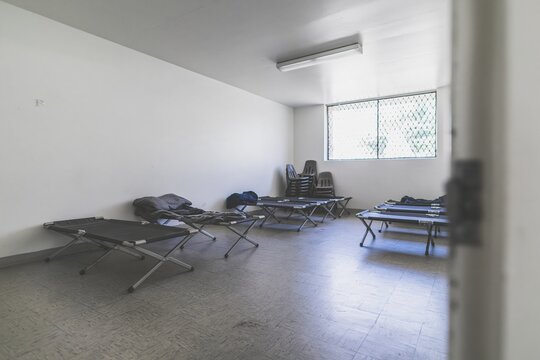 Metal-framed cots in an empty overnight shelter with worn linoleum floors and a gritty, desaturated look for social and documentary projects