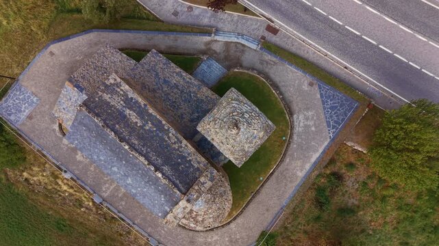 Top-down view of the Romanesque Church of Saint John of Caselles in Canillo, Andorra, with its slate roof, stone bell tower and apse surrounded by grass beside a mountain road