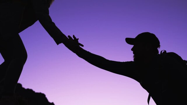 Silhouette of woman helping man climb rocky mountain trail at dusk, reaching out helping hand to pull her companion upward and symbolizing teamwork, support and overcoming challenges