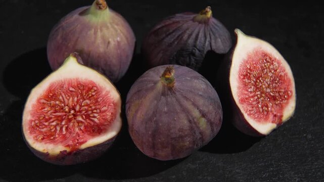 A group of whole purple figs and fig halves slowly rotates on a dark stone surface, showing red flesh and textured skin.