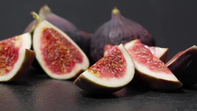 Fresh fig wedges in front of whole purple figs slowly rotate on a dark surface against a dark gray background.