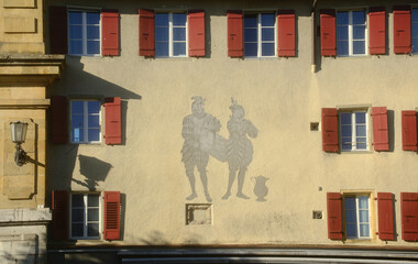 Historic building facade in Morat, Switzerland with red window shutters and a decorative wall painting of two figures carrying a large vessel. © hectorchristiaen