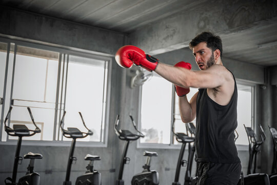 Fit athletic young man throwing a punch wearing red boxing gloves during an intense training session inside a modern gym showcasing dedication to physical fitness and healthy lifestyle.