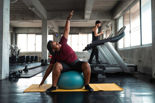 Plus size African American woman stretching back muscles with stability ball on yoga mat in fitness gym. Active healthy lifestyle, flexibility and body positivity concept for everyone.