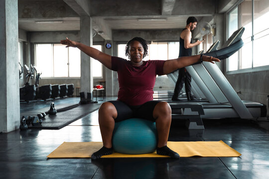 Plus size African American woman stretching back muscles with stability ball on yoga mat in fitness gym. Active healthy lifestyle, flexibility and body positivity concept for everyone.