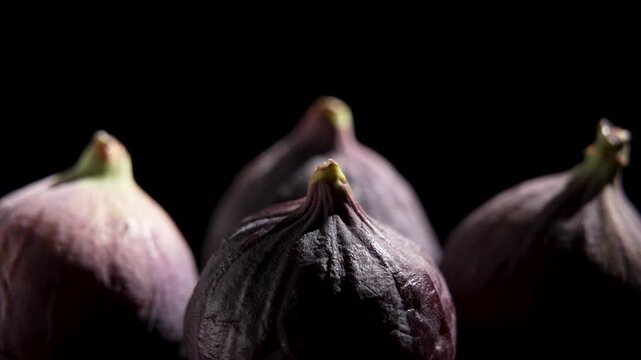 Several whole purple figs with stems slowly rotate in low-key light against a black background, showing ridged skin texture.