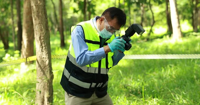 Forensic investigator in protective gear photographs evidence at a taped-off woodland scene, using a camera with flash during a sunny outdoor investigation.