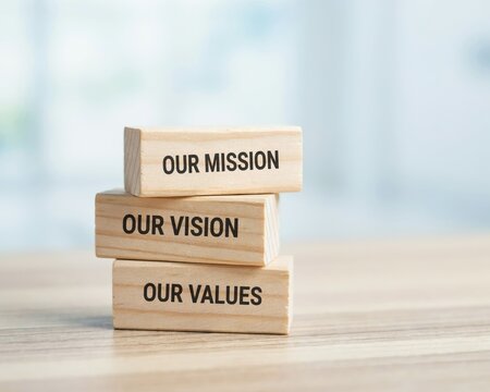 Stack of three wooden blocks on a wooden desk, featuring the engraved text our mission, our vision, and our values in a clear black font style