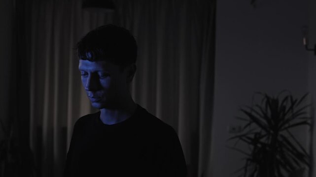 Emotionally drained man standing alone with a downcast gaze, experiencing melancholy and inner emptiness during a difficult life situation, slow camera push-in in a dark evening interior