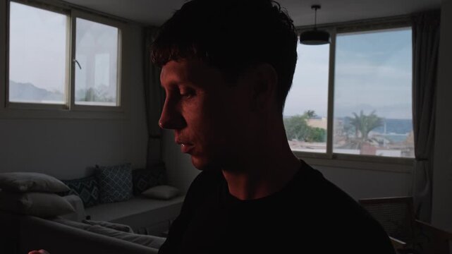 Furious man shouting profanity into his phone during an intense argument at home, then ending the call in anger, with warm light revealing his silhouette against a bright seaside window