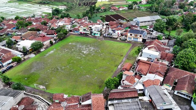 Aerial view of Tanjungsari soccer field surrounded by village houses with red-tiled roofs and lush rice paddies in Sumedang, West Java, Indonesia.