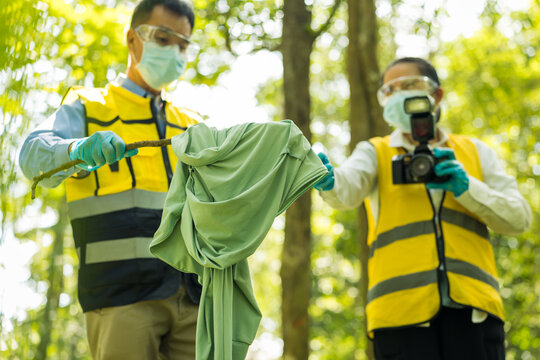 Two masked environmental workers in safety vests collect or document waste in a sunlit forest, holding a green fabric item and camera during an outdoor cleanup inspection.