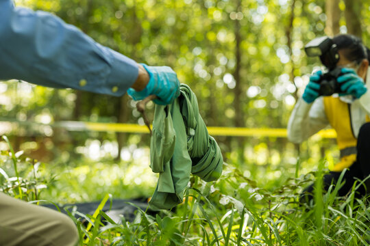 Forensic investigators examine clothing evidence in a wooded crime scene behind yellow police tape, with one investigator photographing the area and another wearing protective gloves.
