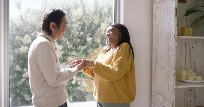 Senior Asian man taking African-American woman's hand proposing by placing ring on finger by window