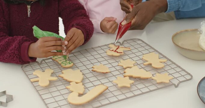 Left child and adult are squeezing piping bags while decorating cookies on counter for christmas