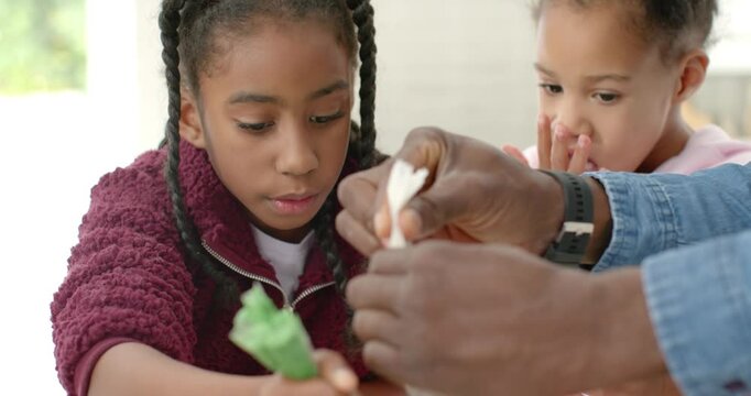 Adult hands guiding African American schoolgirls piping christmas cookies with piping bag at table