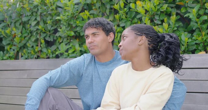 Couple laughing and talking, laughter leading to leaning on bench by green hedge showing affection