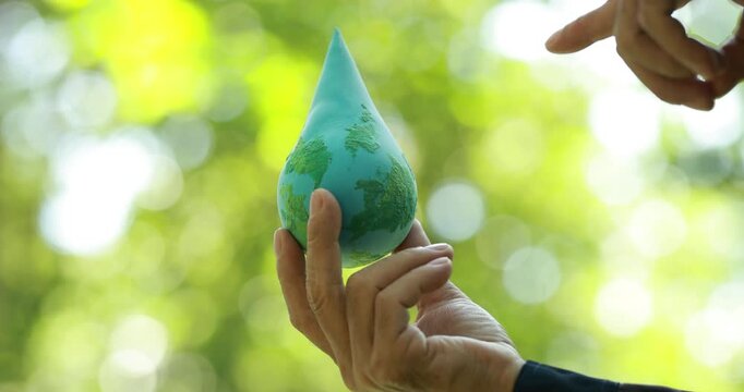 Hands hold a blue water drop shaped like a globe outdoors, symbolizing water conservation, environmental care, sustainability, and global responsibility against a soft green nature background.