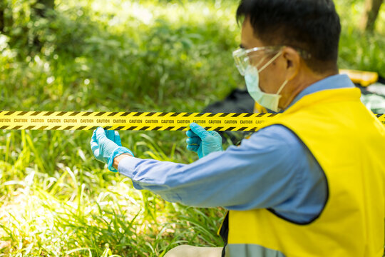 A masked field investigator in protective clothing and gloves works behind caution tape in a sunny green outdoor area, examining or collecting evidence.