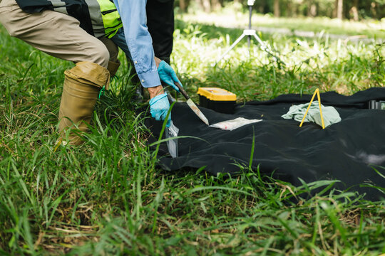 Field workers in protective clothing and gloves use hand tools beside a black ground sheet in a grassy park during an outdoor environmental inspection or soil survey.
