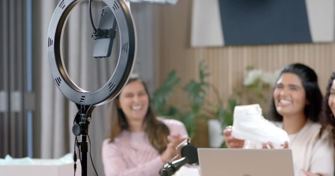 Diverse female co-hosts, middle lifting sneaker while others clapping and reviewing near ring light