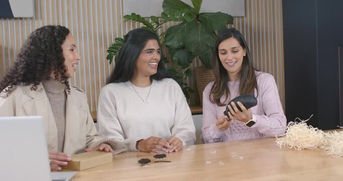Diverse female coworkers pulling black zippered pouch from box at studio table, examining contents