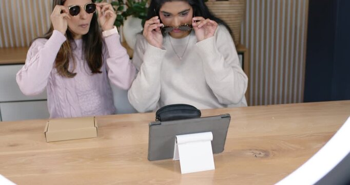 Diverse women friends spotting sunglasses, testing via tablet stand; third woman joining