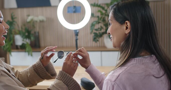 Diverse female friends: pink-sweater holding shades, beige coat trying on at table with ring light