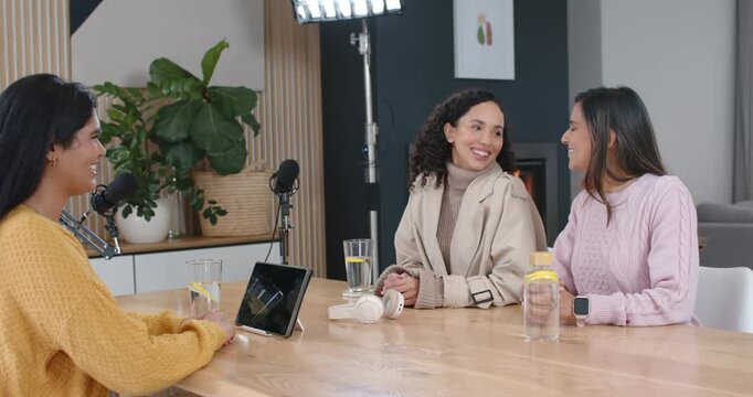 Diverse female colleagues recording podcast in studio with mics, Indian host tapping tablet