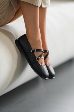 Close-up of woman legs and feet wearing black Mary Jane shoes with buckled ankle straps on sofa in warm indoor light with soft neutral background (warm indoor light with soft neutral background)