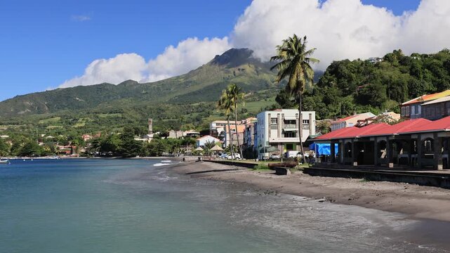 Mount Pelee, an active volcano, rising prominently above the lush green tropical forest and palm trees of Martinique, French Antilles, under a bright blue sky with white clouds