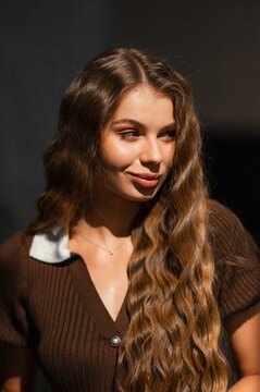 Smiling young woman with long brown wavy hair wearing brown knit top in soft studio lighting warm inviting portrait with closeup focus on face and expression, friendly mood