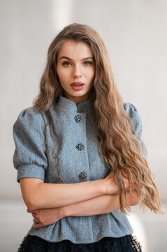 Portrait of young woman with long wavy hair wearing gray textured coat, arms crossed indoors with gentle lighting against soft neutral background, 34-year-old vibe