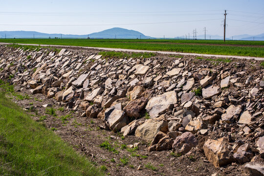 Stone-lined drainage channel for flood protection and erosion control in a rural agricultural landscape with green fields, hills, and utility infrastructure.