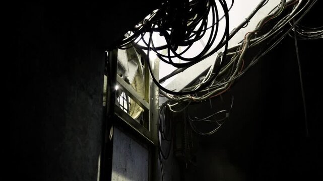 Dense cable nest inside service ceiling, frayed wires and metal conduits tangled near opening, light leak accentuates industrial complexity and maintenance