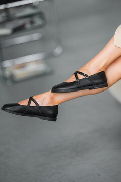 Close-up of black double-strap ballet flats on extended leg beside parked car in indoor parking garage, beige skirt and minimalist urban fashion under modern concrete lighting