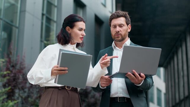 Two serious Caucasian business workers talking looking in laptop comparing data in online software and documents in city. Mature businessman holding computer businesswoman hold folder talk outdoors