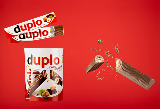 Ferrero Duplo chocolate-covered hazelnut wafer bars on a red background, featuring a ten-bar multipack, a wrapped stick, a partly unwrapped bar, and a broken wafer bar with floating crumbs.