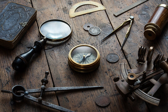 Antique navigation and exploration still life centered around an old compass on a rustic wooden surface, surrounded by an astrolabe, dividers, antique book, coins, spyglass, and magnifying glass