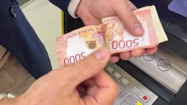 Close up man hands count a thick stack of Russian ruble banknotes, five thousand beside bank automated teller machine, showing red and pink currency details. Withdrawing cash from an ATM. footage