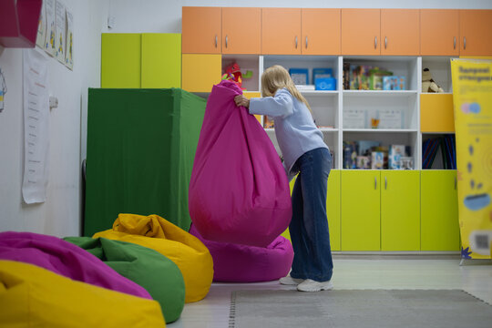 Child lifts a large pink bean bag in a colorful playroom with shelves and other bean bags