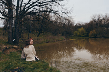 Fototapeta premium Woman sits by a tranquil lake in a park wearing a cozy sweater, reflecting on a quiet day. Soft light and muted tones create a calm mood, highlighting nature and autumn ambiance.